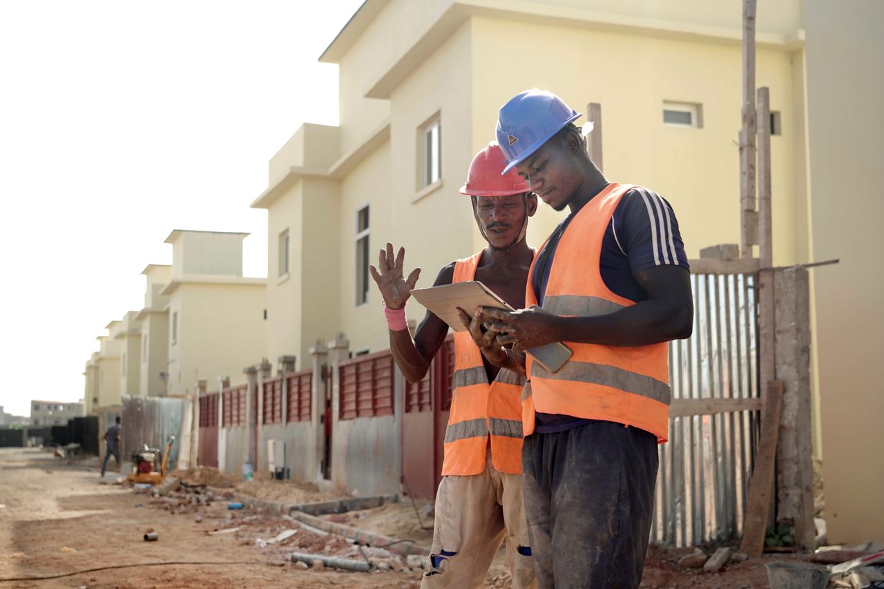 our-services-02 Two construction workers in helmets and vests review blueprints on a site.