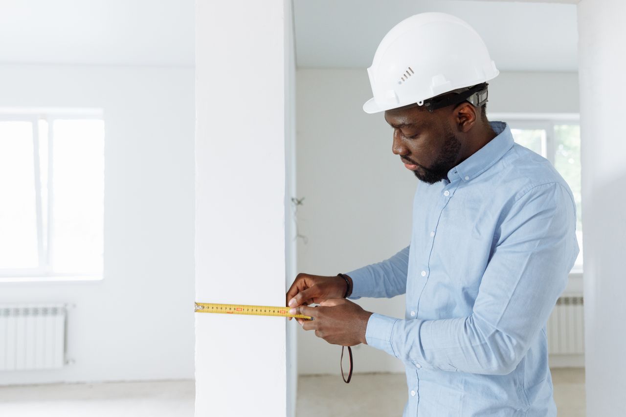 our-services-03 Construction worker measuring a wall indoors, wearing a white hard hat and blue shirt.
